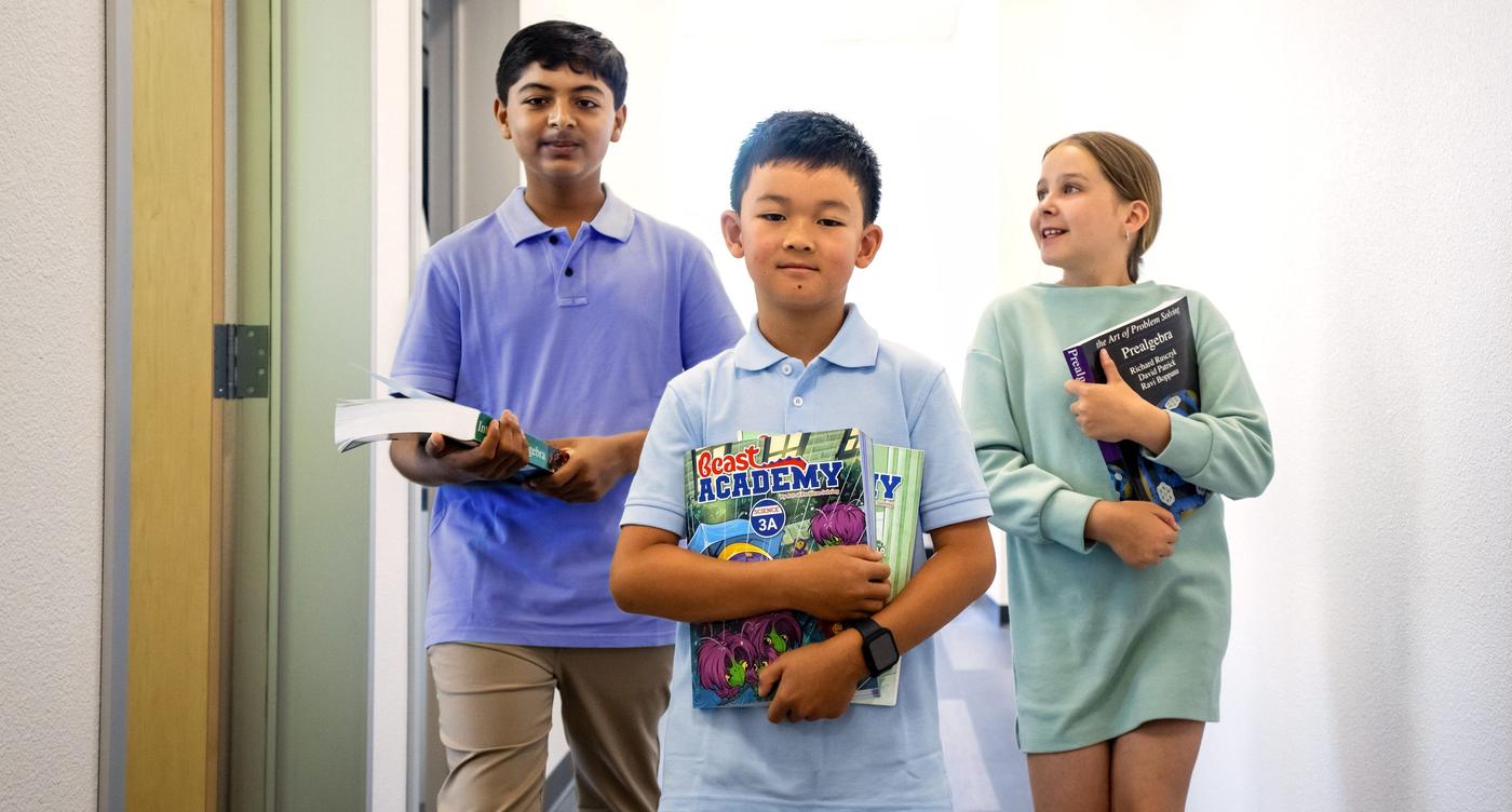 Three AoPS Academy math students walk through hallway holding Beast Academy and AoPS books 